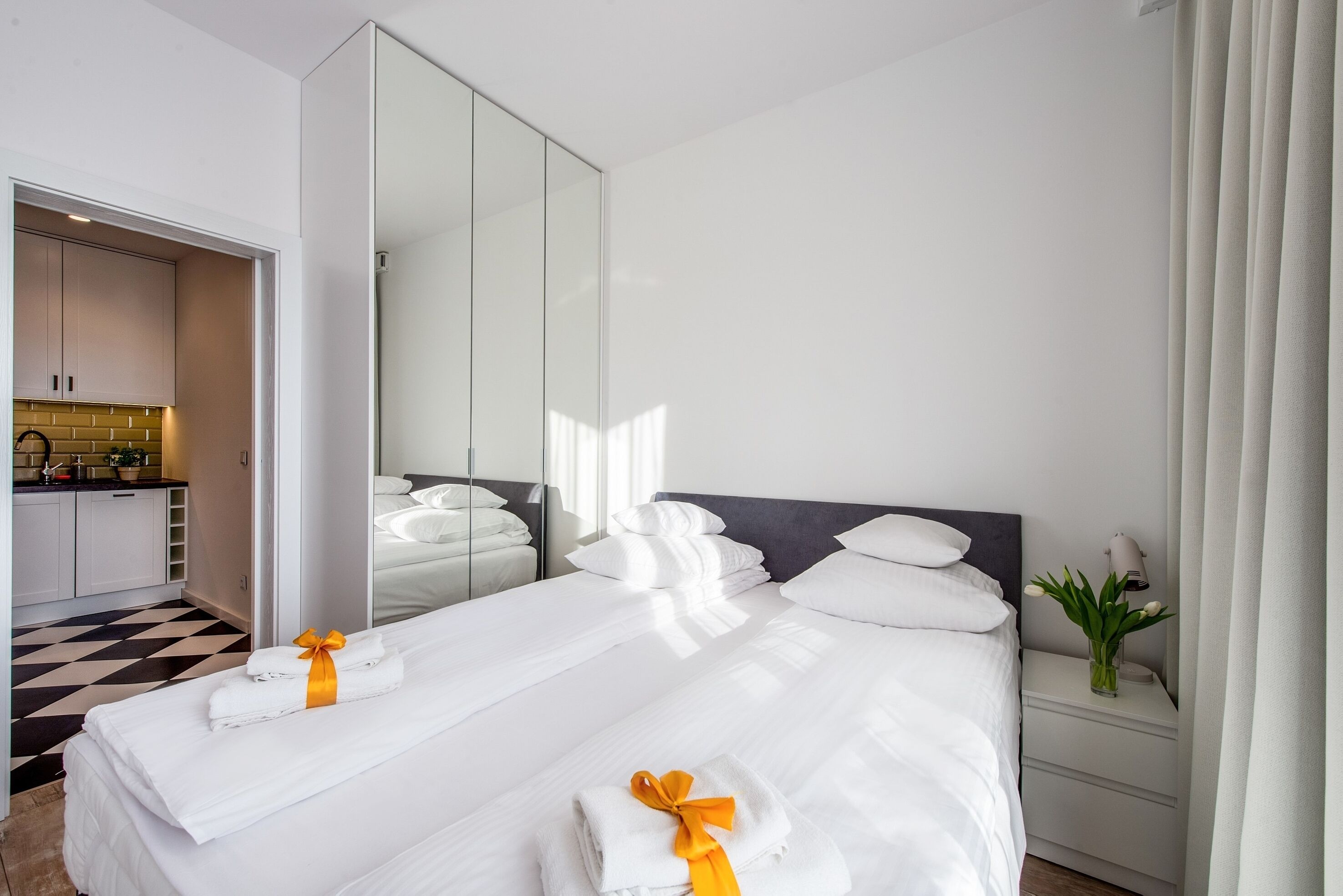 A hotel room with two beds, a large mirror, a nightstand with a lamp, and a view of the kitchen.
