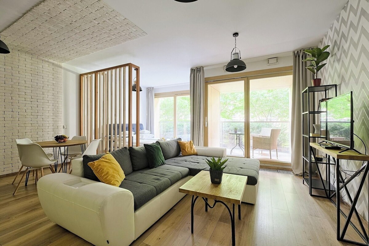 A modern living room with a sectional sofa, a coffee table, a dining area with chairs, a bookshelf, and a view of the outdoors.