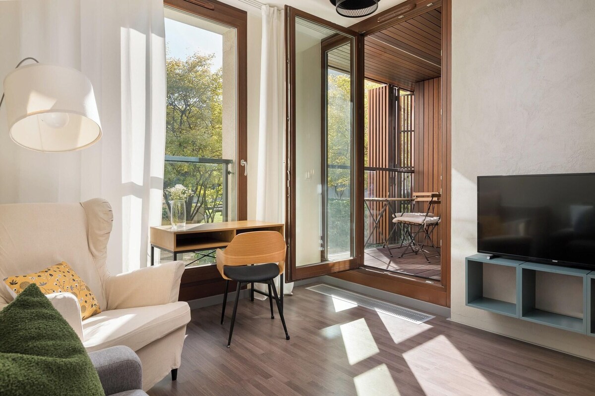 A modern living room with a sofa, a desk with a chair, a television, and a balcony with outdoor furniture.