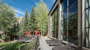 A deck with lounge chairs and a red umbrella, surrounded by trees and a building with large windows. - Remodeled and Oversized 2-bedroom Condo in West Keystone With Mountain Views, Pool, hot tub & Shuttle to Slopes! (Keystone)