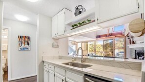 A modern kitchen with white cabinetry, a stainless steel sink, and a dishwasher. There is a window with a view of the outdoors, and a shelf with decorative items above the sink. - Remodeled and Oversized 2-bedroom Condo in West Keystone With Mountain Views, Pool, hot tub & Shuttle to Slopes! (Keystone)