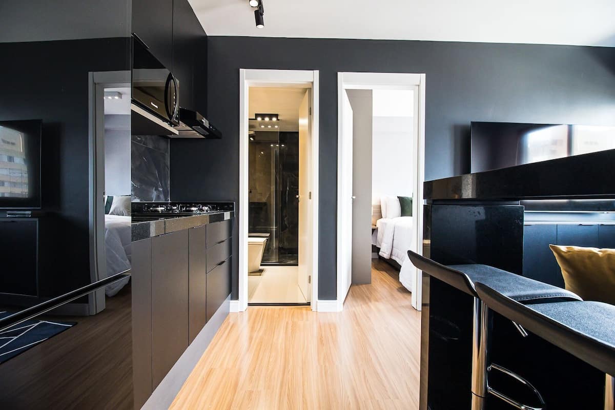 A modern kitchen with dark cabinetry, a wooden floor, and a view into a bedroom.