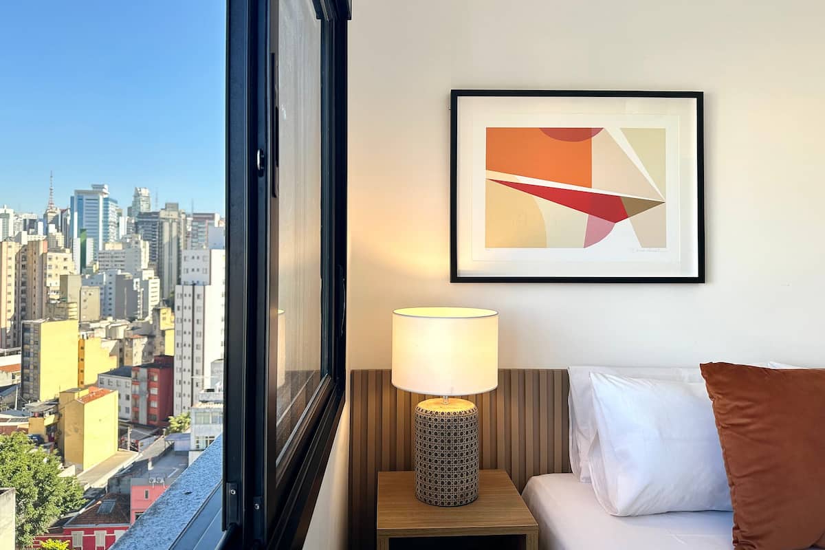 A hotel room with a city view, a framed abstract painting, a bedside lamp, and a bed with pillows.
