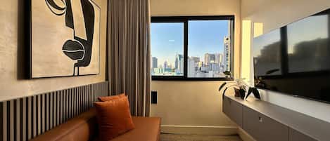 A modern hotel room with a city view, a large flat-screen TV, a sofa with a cushion, and a framed abstract artwork on the wall.