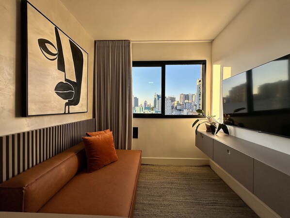 A modern hotel room with a city view, a large flat-screen TV, a sofa with a cushion, and a framed abstract artwork on the wall.