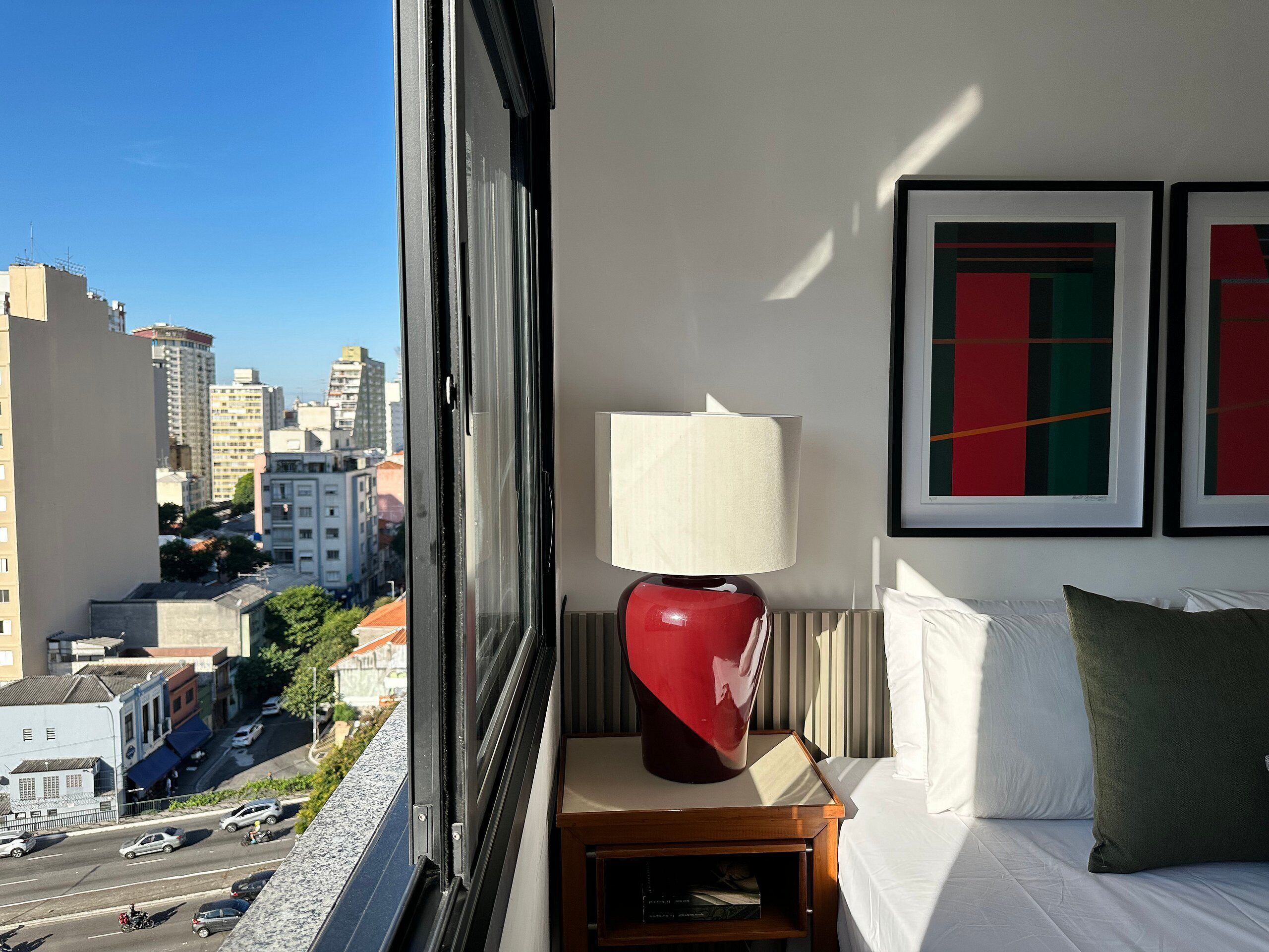 Une chambre d’hôtel avec un lit, une table de chevet, une lampe et deux œuvres d’art encadrées. On peut voir un paysage urbain à travers la fenêtre.