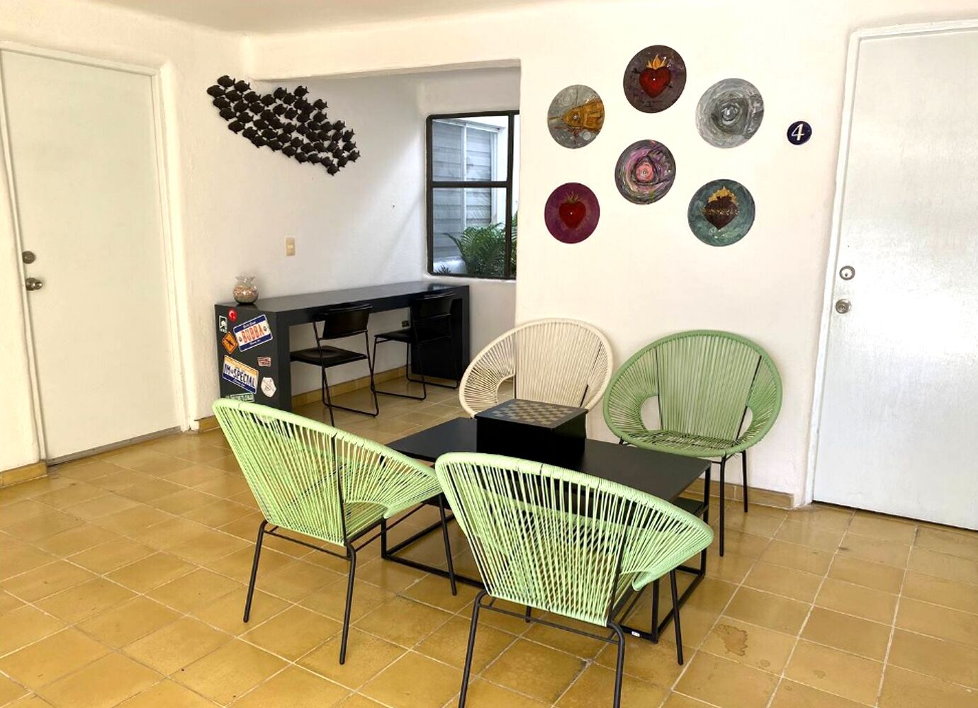 Uma sala de jantar com uma mesa preta e quatro cadeiras verdes. Há pratos decorativos na parede e uma lousa com adesivos.