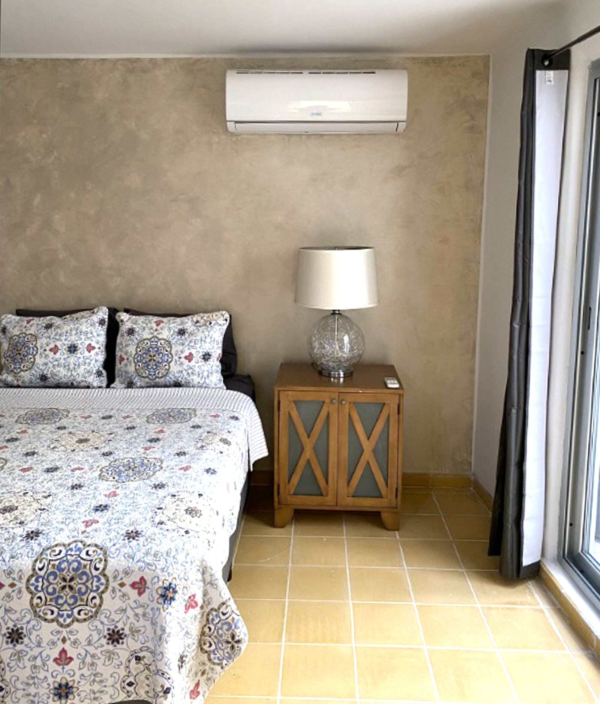 Ein Schlafzimmer mit Bett, gemusterten Textilien, einer Nachttischlampe, einem Holfnachtkästchen und einer Wandklimaanlage.