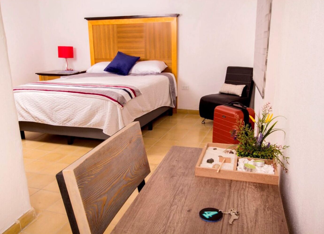 A hotel room with a bed, a desk, a chair, a red suitcase, and a small table with a plant and a keychain.
