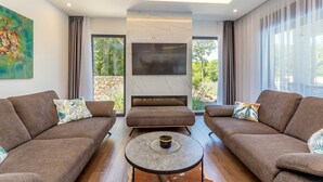 Un salon moderne avec deux canapés, une table basse, un téléviseur à écran plat fixé au mur et de grandes fenêtres donnant sur un paysage verdoyant.