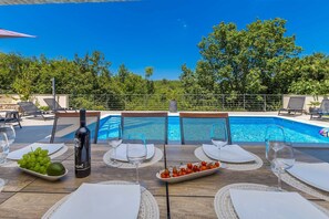 Uma área de refeições à beira da piscina com vista para uma piscina azul cristalina, cercada por vegetação exuberante.