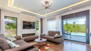 Un salon moderne avec un ensemble de canapés, une table basse et un téléviseur à écran plat fixé au mur. Une grande fenêtre offre une vue sur la verdure à l’extérieur.