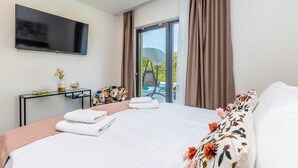 Une chambre d’hôtel moderne avec un grand lit, un téléviseur à écran plat fixé au mur, un petit bureau agrémenté d’un vase de fleurs et une vue sur la piscine, accessible par une porte-fenêtre coulissante.