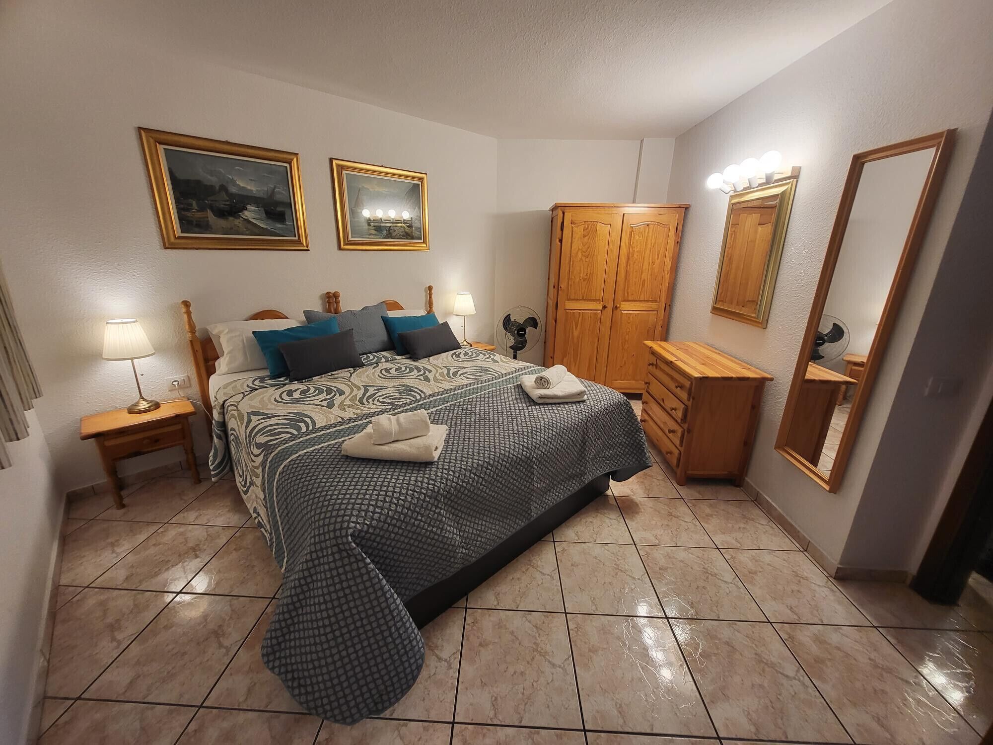 A bedroom with a bed, a wooden wardrobe, a bedside table with a lamp, a dresser, and a large mirror.