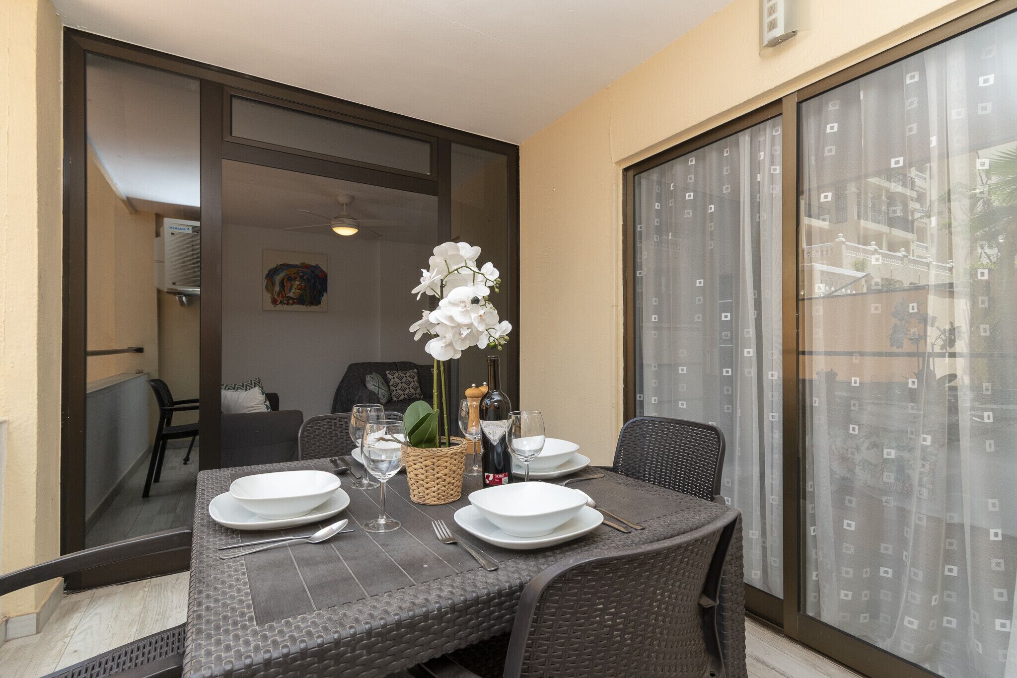 Een eethoek met een tafel gedekt voor twee personen, een rieten stoel, een rieten bank, een witte orchidee en een fles wijn.