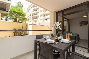 Un balcon avec une table à manger pour deux personnes, un pot de fleurs et une vue sur un immeuble à plusieurs étages.
