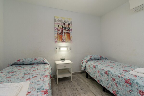 A small, clean room with two single beds, a bedside table with a lamp, and a wall-mounted light fixture.