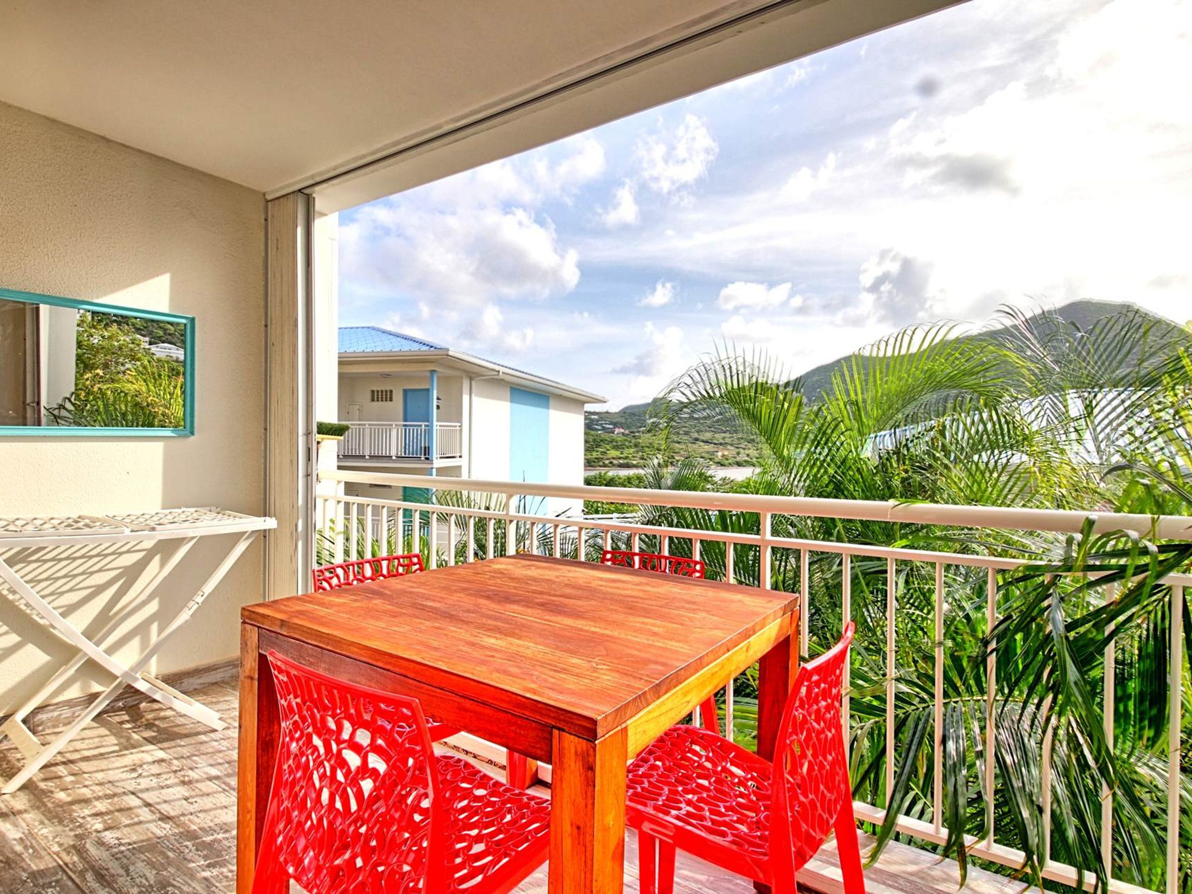Un balcón con una mesa de madera y sillas rojas, con vistas a una zona residencial con casas y palmeras.