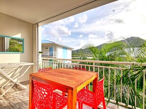 A balcony with a wooden table and red chairs, overlooking a residential area with houses and palm trees.