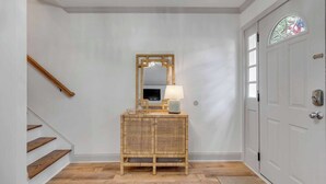Un couloir avec un escalier en bois, une console en osier agrémentée d’un miroir et d’une lampe, et une porte blanche ornée d’un vitrail.