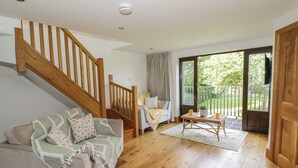 A spacious living room with a wooden staircase, a sofa with a throw blanket, a coffee table, and a large sliding glass door leading to a garden. - Hillcroft (Kenilworth)