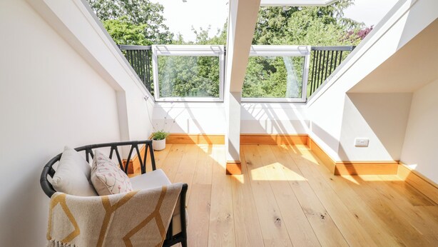 A room with a wooden floor, a black chair with cushions, a white sofa, and a patterned throw blanket. There are two skylights providing natural light and a view of greenery outside. - Hillcroft (Kenilworth)