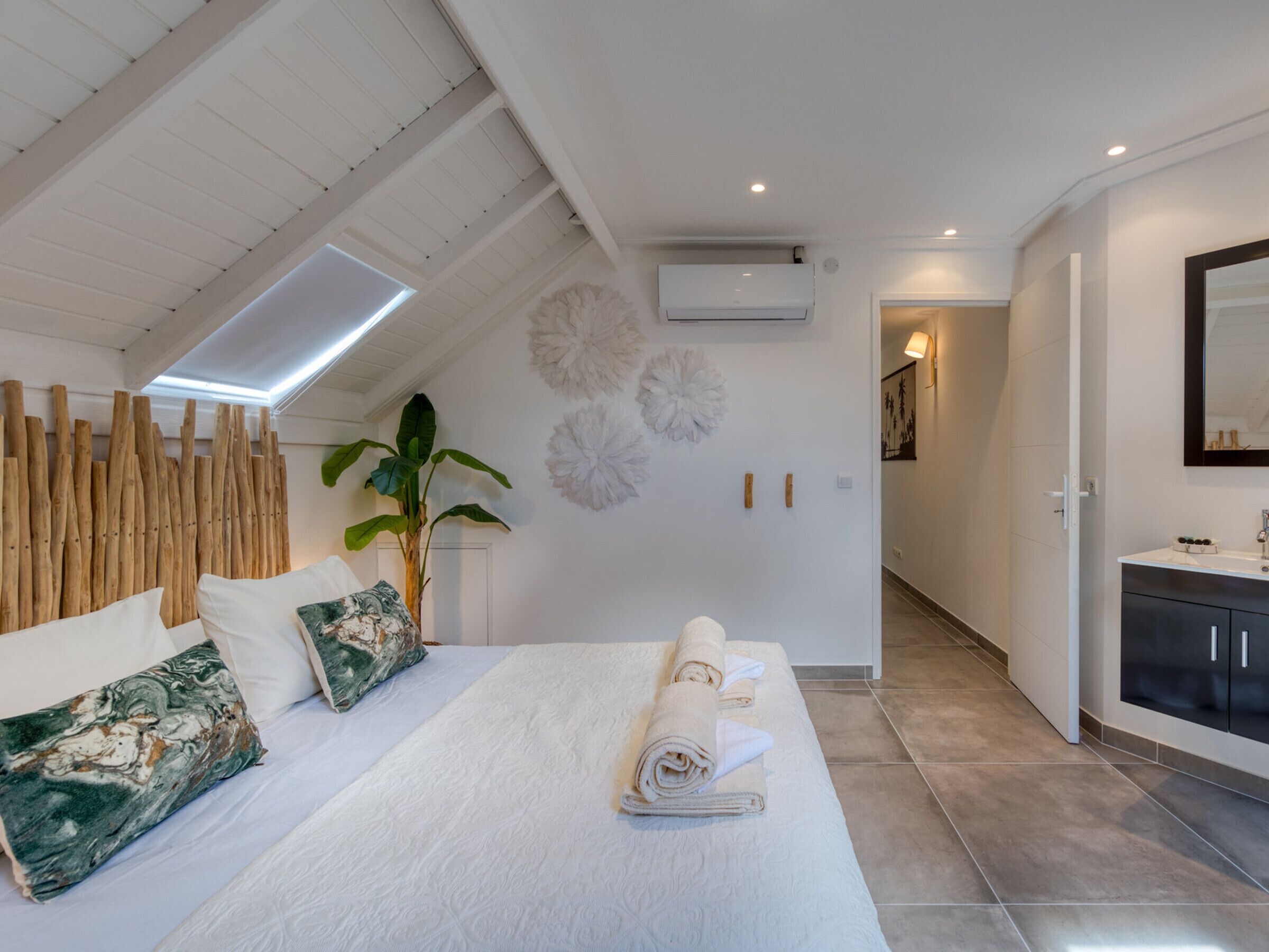 A modern bedroom with a large bed, a potted plant, and a bathroom with a mirror and sink.