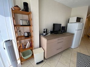Uma kitchenette compacta com micro-ondas, torradeira e máquina de café. Há um pequeno contentor de lixo e uma prateleira com livros e utensílios de cozinha.