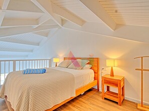 A bedroom with a sloped ceiling, a wooden bed frame, a bedside table with a lamp, and a view of the outdoors.