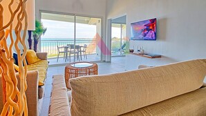 A modern living room with a large sofa, a television, and a view of the ocean through a sliding glass door.
