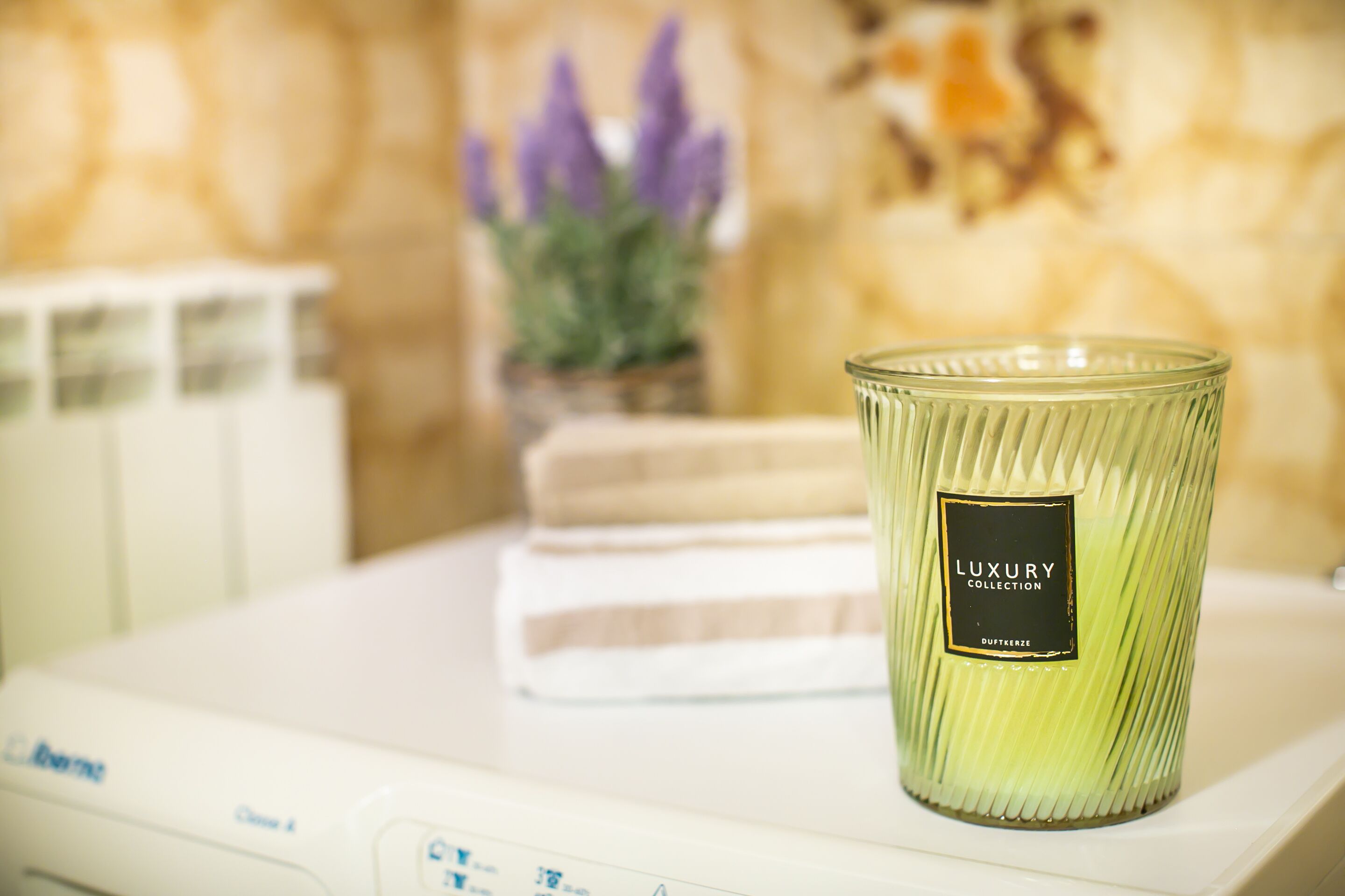 Une bougie verte portant l’étiquette « Collection de luxe » est posée sur une table blanche, avec une serviette et une plante en pot en arrière-plan.