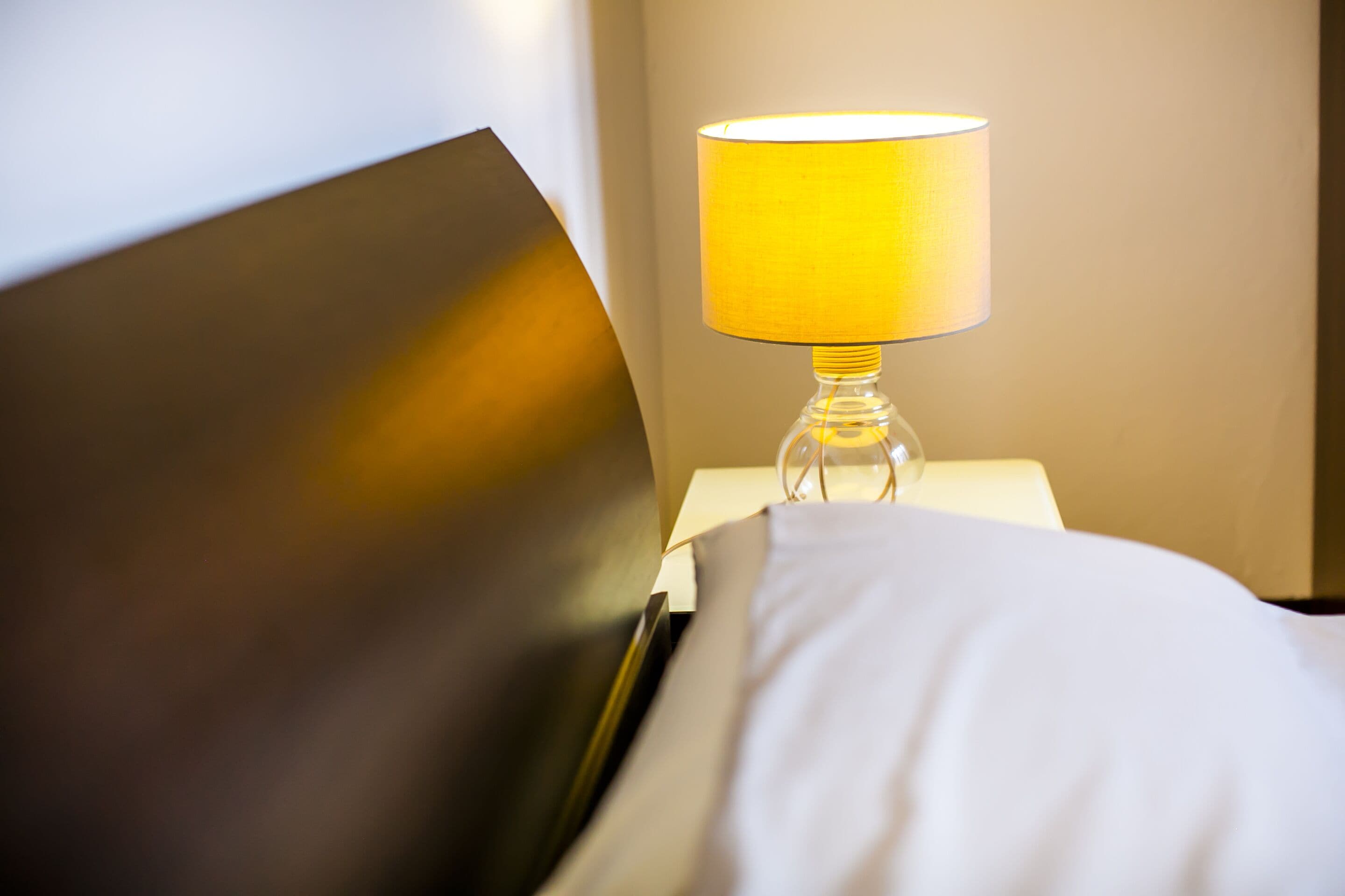 A bed with a white comforter and a yellow lamp on a bedside table.