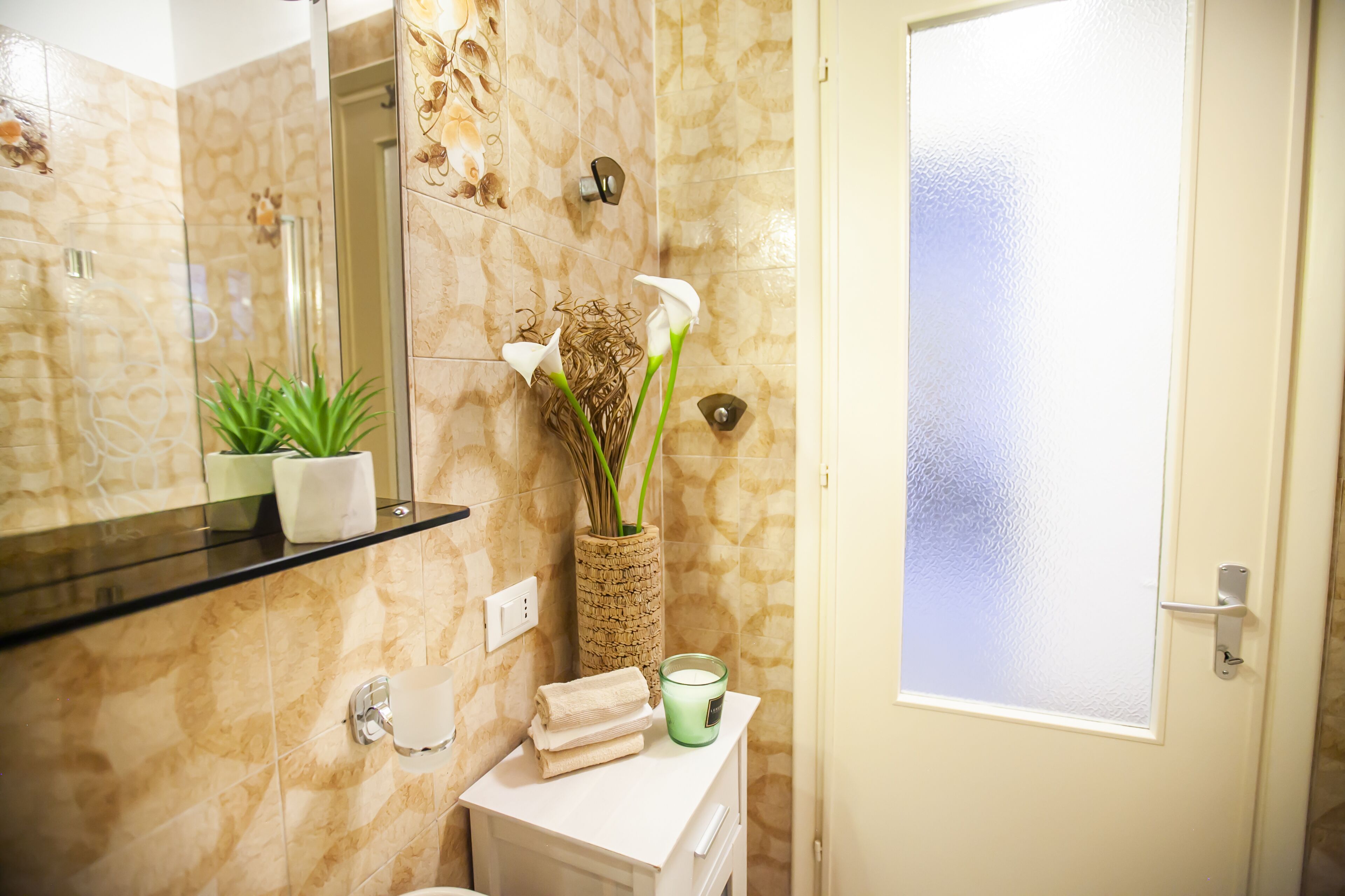 A bathroom with a marble wall, a glass shower enclosure, a white sink, a mirror, a vase with flowers, and a frosted glass door.