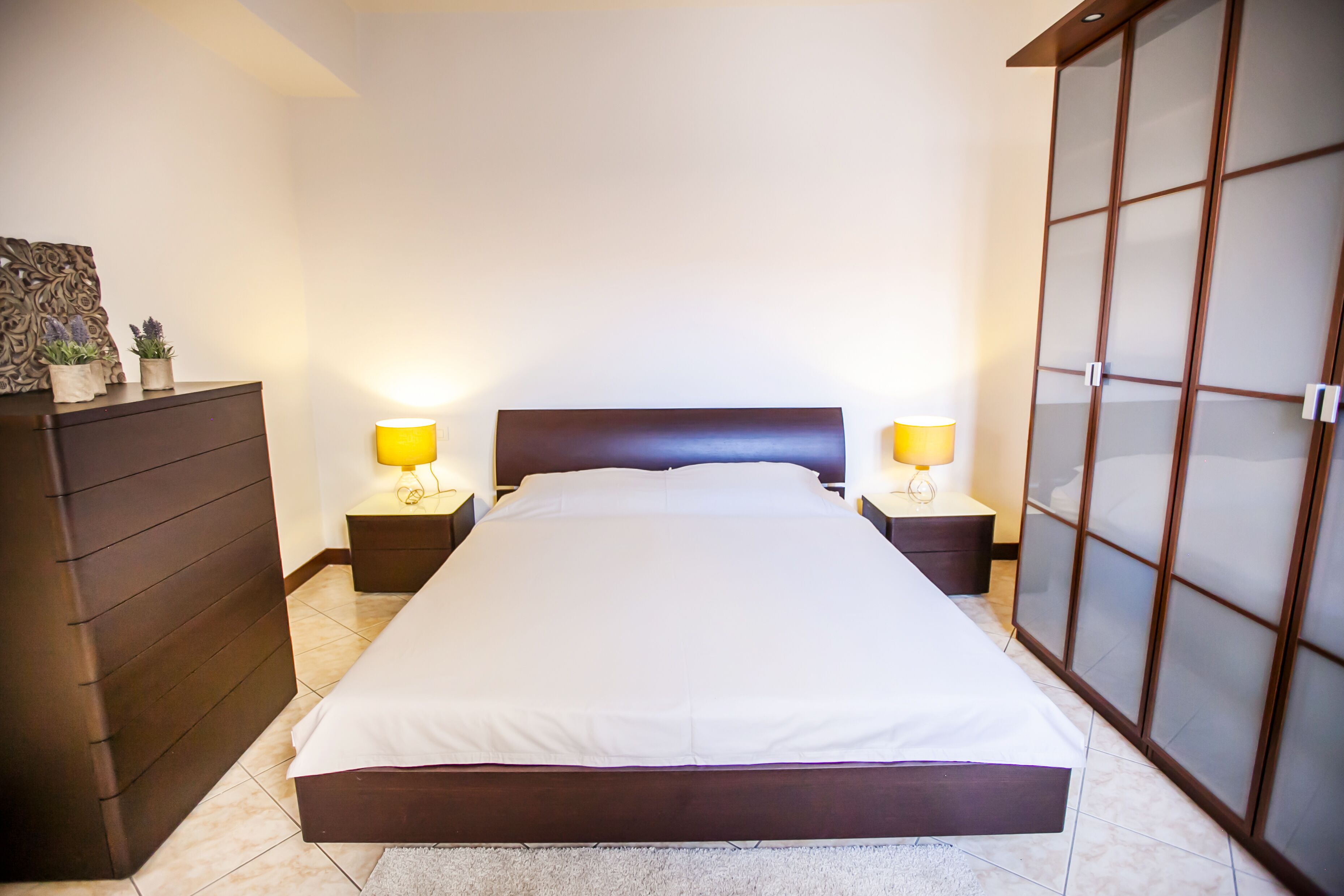 A neatly arranged bedroom with a large bed, two bedside tables with lamps, a dresser, and a sliding glass door closet.