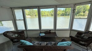 A room with wicker furniture, a table, and large windows overlooking a lake.