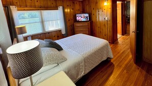 A wooden-paneled bedroom with a bed, a TV, a ceiling fan, and a window with curtains.