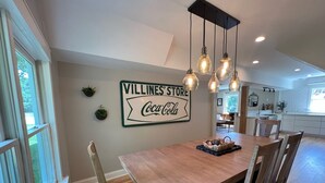 Un área de comedor con una mesa y sillas de madera, un letrero que dice "Villines' Store Coca-Cola" y lámparas colgantes.