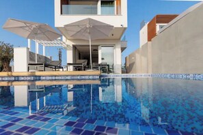Un espace piscine moderne avec un sol en mosaïque bleu clair, deux parasols et un bâtiment contemporain en arrière-plan.