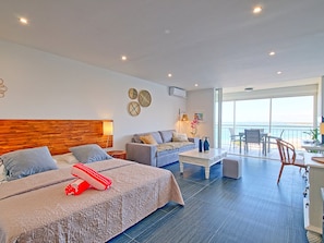 A modern hotel room with a large bed, a sofa, a dining area, and a balcony with a view.