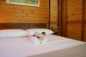Una cama bien hecha con ropa blanca y toallas enrolladas, un cabecero de madera y un pequeño florero con flores sobre una mesita de noche.