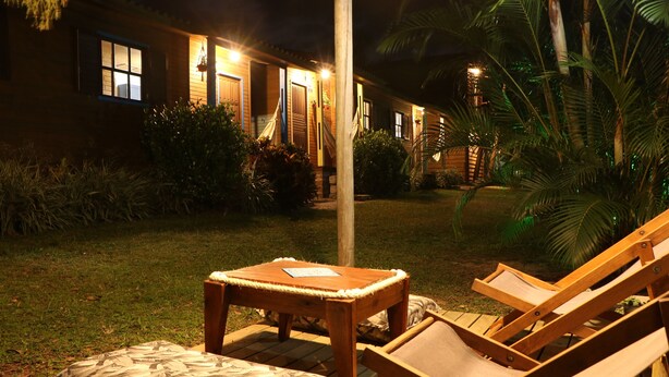 Una terraza de madera con una mesa y una silla de salón, rodeada de abundante follaje y un edificio con ventanas iluminadas.