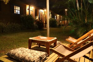 A wooden deck with a table and a lounge chair, surrounded by lush greenery and a building with illuminated windows. - Chalés Vale da Lua Praia do Rosa (Imbituba)