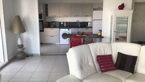 A modern living room with a sofa, a lamp, and a kitchen in the background. - Appt Standing Face à la Mer (Fréjus)