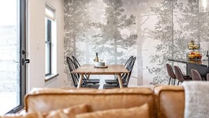 A dining area with a wooden table and chairs, a wall with a tree pattern, and a window with a view.