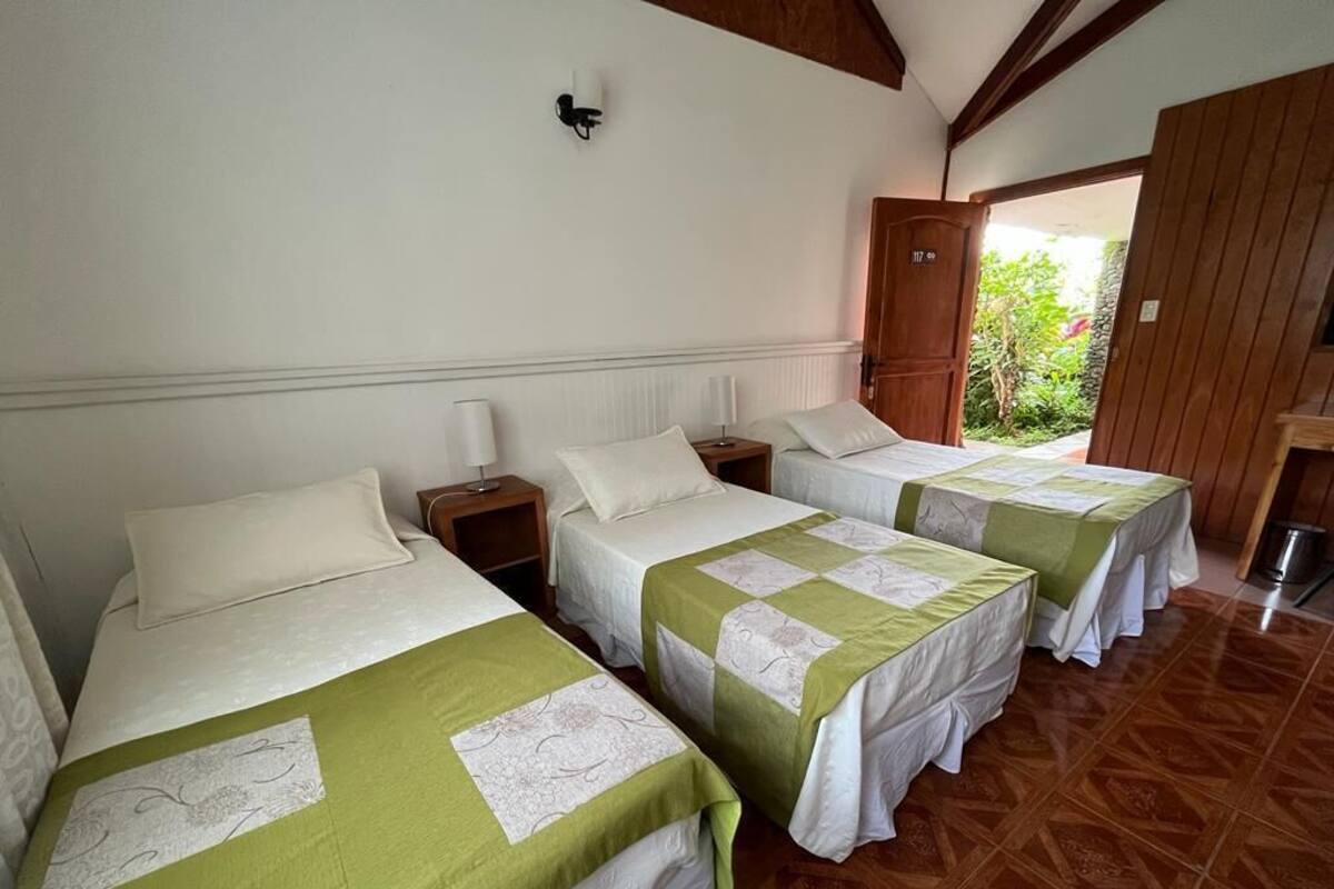 A room with three beds, each with a green and white bedspread, wooden headboards, and bedside tables with lamps.