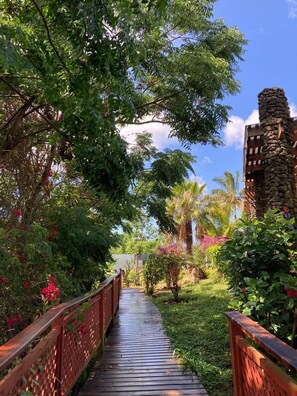 A wooden pathway leading through a lush garden with various plants and a stone structure. - Hare Rapa Nui by Chez Joseph (Hanga Roa)