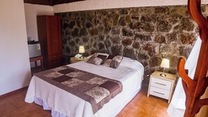 A bedroom with a stone wall, a bed with a patterned bedspread, two bedside tables with lamps, and a wooden chair.