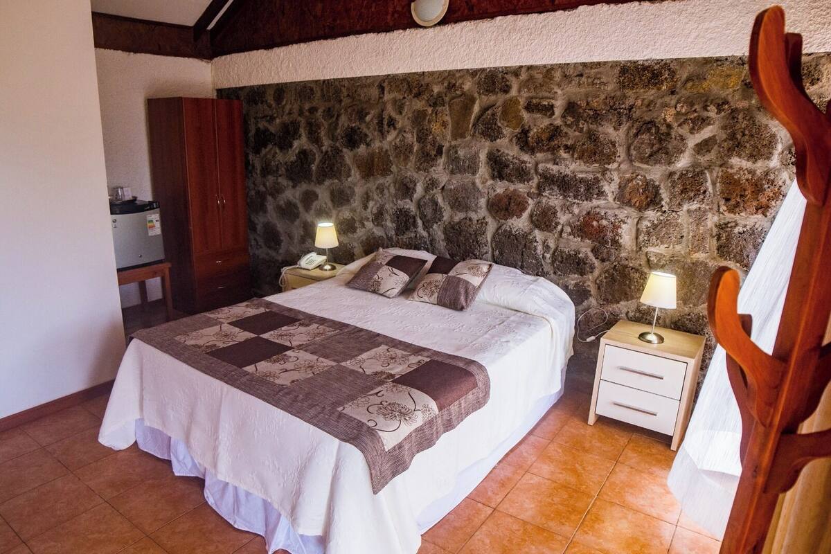 A bedroom with a stone wall, a bed with a patterned bedspread, two bedside tables with lamps, and a wooden chair.