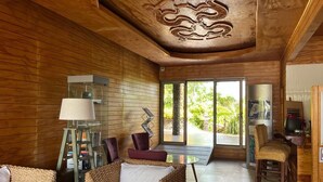 A room with wooden walls, a ceiling with a carved design, wicker furniture, a glass table, and a view of greenery outside.