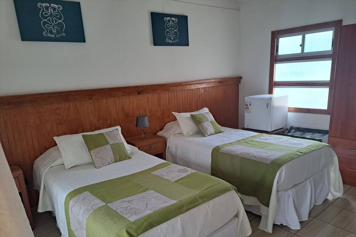 A hotel room with two beds, wooden headboard, bedside tables with lamps, a small refrigerator, and a window with blinds.